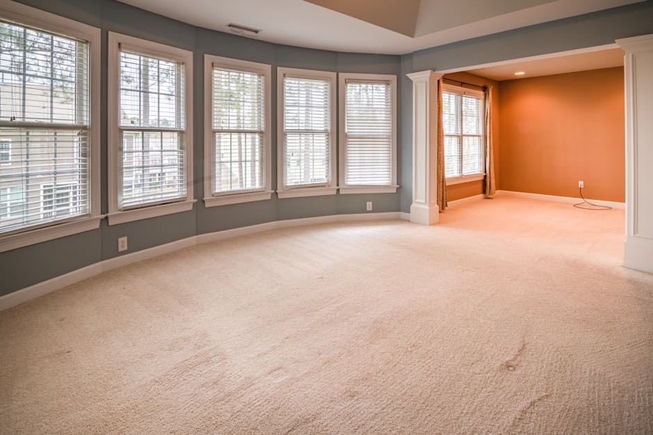 An image of a spacious living room with light beige carpeting that appears clean and well-maintained. The room is illuminated by natural light coming through multiple white-framed windows fitted with