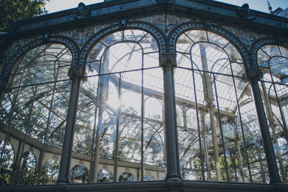 An ornate Victorian-style glass conservatory featuring tall, curved glass panels supported by decorative metal columns with intricate patterns. The glass surfaces are clean and clear, allowing sunligh